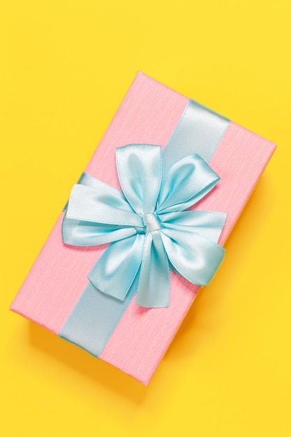 Pink gift box tied with blue ribbon with bow at the top on yellow background.