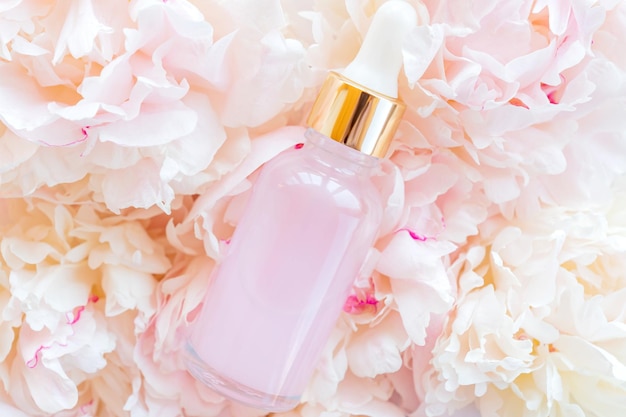 A pink glass bottle with a serum essential oil or other cosmetic product against a background of pink peonies Natural organic cosmetic packaging skin care concept