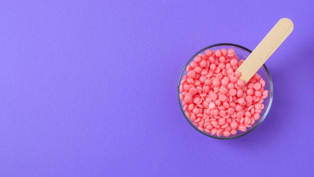 Pink granules of wax for depilation in a glass bowl on a lilac background Wooden spatula Banner