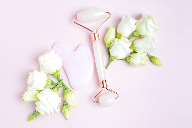 Pink Gua Sha facial massage and face care tools Rose Quartz jade roller on pink background with eustoma flowers composition Anti age lifting and toning treatment at home top view Copy space