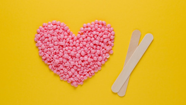 Pink heartshaped wax granules for depilation and wooden spatulas on a yellow background Epilation