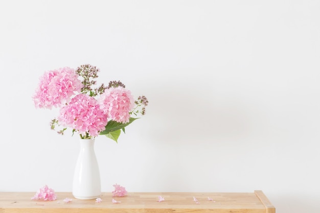 Pink hydrangea in white vase on wooden shelf on background white wall