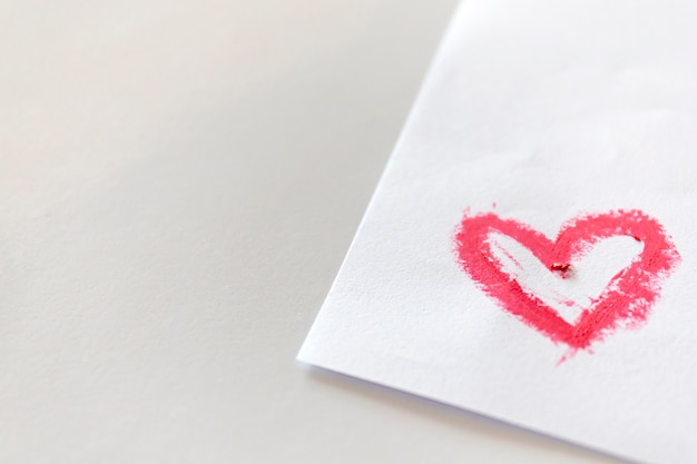 Pink lipstick smeared in heart shape on white paper on white table.