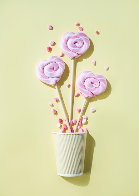 Pink lollipops in the shape of hearts at paper cup with color sugar on a yellow background Top view and copy space Vertical orientation