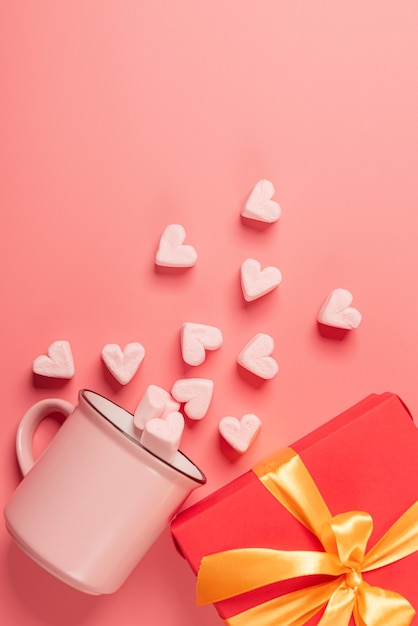 A pink mug lies on its side with marshmallows sprinkled from it in the form of hearts and gift