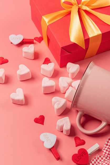 A pink mug lies on its side with marshmallows sprinkled from it in the form of hearts and gift