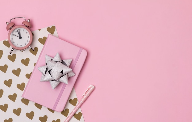 Pink notepad with alarm clock, bow and pen on creative background