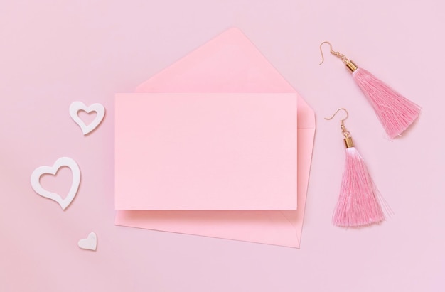 Pink paper card and envelope, hearts and tassel earrings on pink top view. Minimal scene with blank card mockup. Valentines day, educational and girly concept, space for text.