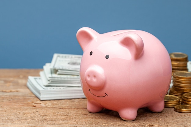 Pink piggy bank and money with banknotes and coins on a wooden table and blue background.