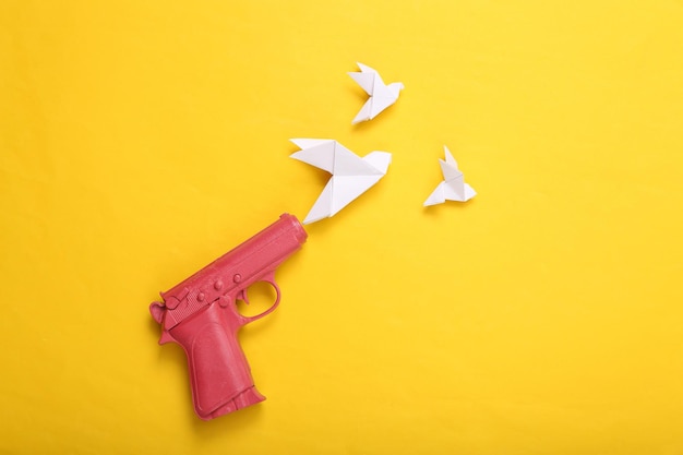 Pink pistol gun shooting origami doves on yellow background No war Creative minimalistic layout Flat lay Top view