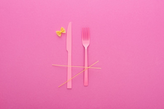 Photo pink plastic knife and fork spaghetti and uncooked farfalle pasta isolated on pink