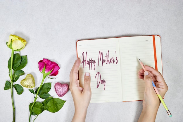 Photo pink and yellow roses flowers and glitters hearts woman hand writing happy mothers day on notebook