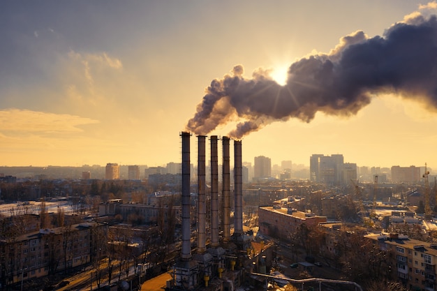 Pipes of an industrial factory with black smoke covering the sun during the yellow sunset in winter in the city