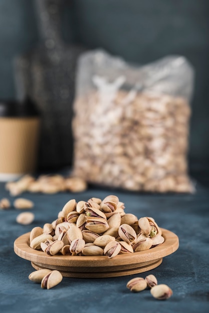 Pistachios on a wooden plate and in a package with a coffee cup on a dark blue background. A natural product for the diet. Side view with a copy space. Vertical orientation.