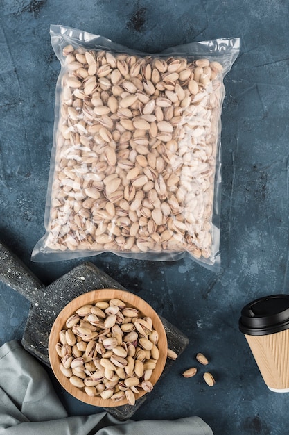 Pistachios on a wooden plate and in a package with a coffee cup on a dark blue background. A natural product for the diet. Top view with a copy space. Vertical orientation.