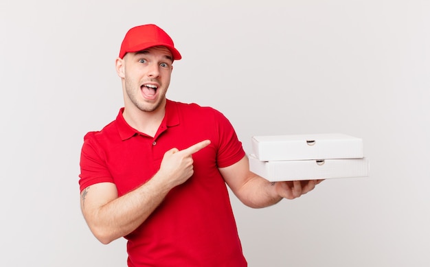 Pizza deliver man looking excited and surprised pointing to the side and upwards to copy space