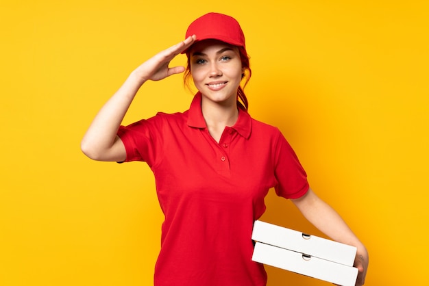 Pizza delivery girl holding a pizza over isolated wall saluting with hand with happy expression