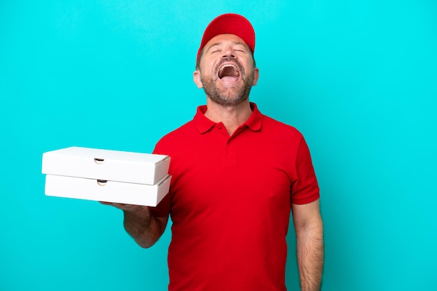 Pizza delivery man with work uniform picking up pizza boxes isolated on blue background laughing