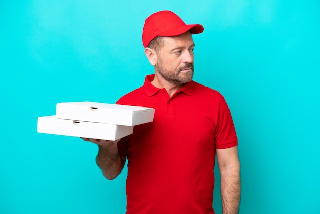 Pizza delivery man with work uniform picking up pizza boxes isolated on blue background looking to the side