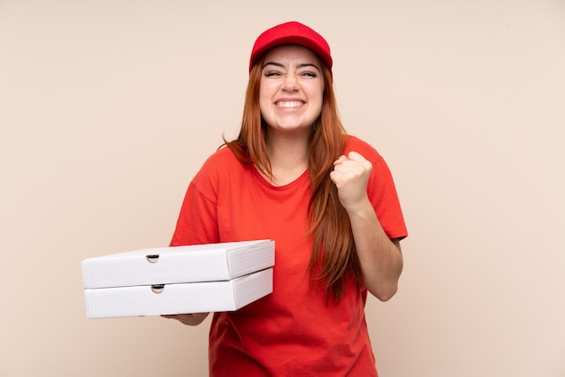 Pizza delivery teenager girl holding a pizza over isolated wall celebrating a victory