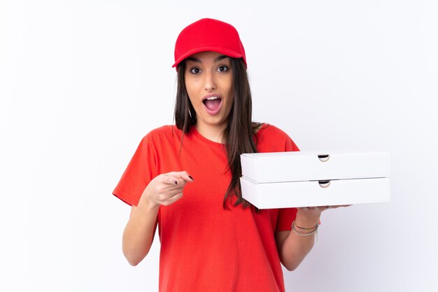 Pizza delivery woman holding a pizza over white wall surprised and pointing front