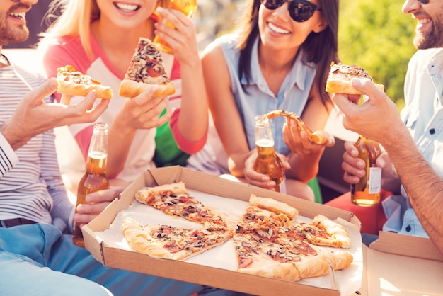 Pizza time! Close-up of four young cheerful people eating pizza and drinking beer outdoors