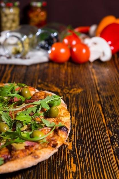 Pizza with arugula and olives on wooden table, tomatoes, mushrooms and other vegetables in the background