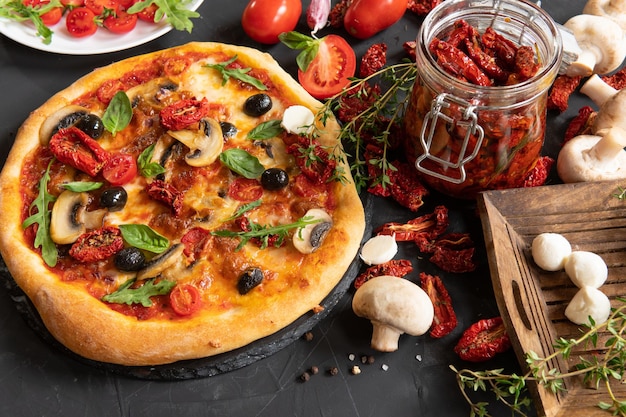 Pizza with vegetables and dried tomatoes on a dark background in the kitchen.