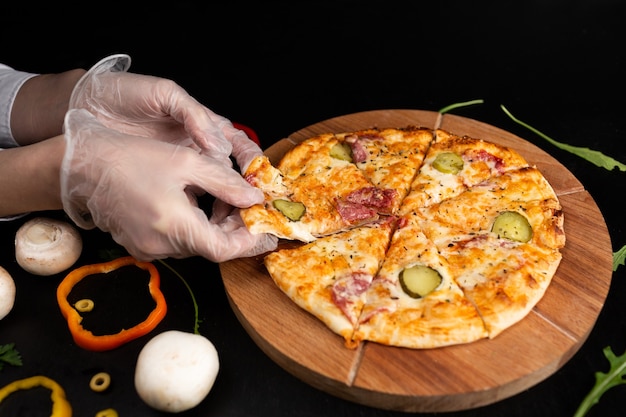 Pizza on a wooden stand on a black surface Gloved hands take a slice of pizza