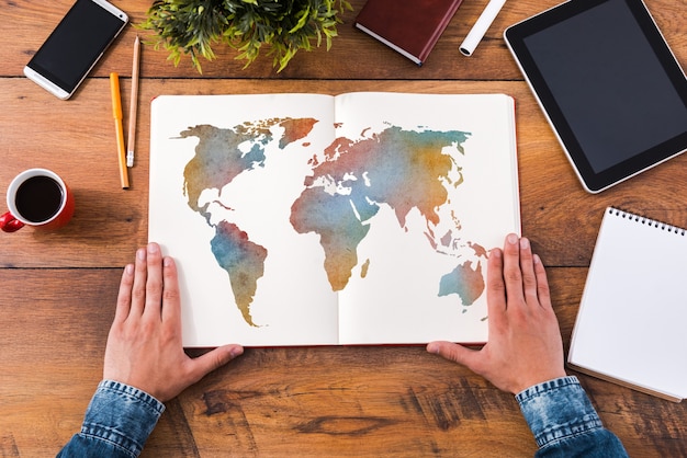 Planning his journey. Top view close-up image of man holding hands on his notebook with colorful map on it while sitting at the wooden desk