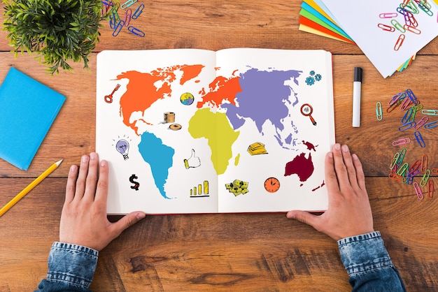 Planning his journey. Top view close-up image of man holding hands on his notebook with colorful map on it while sitting at the wooden desk