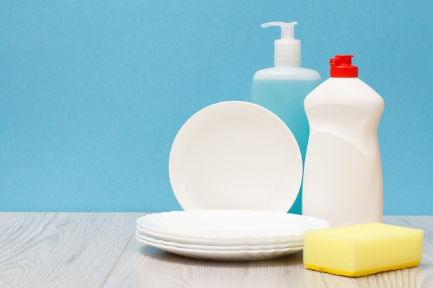 Plastic bottles of dishwashing liquid, glass and tile cleaner, clean plates and sponge on blue background. Washing and cleaning concept.