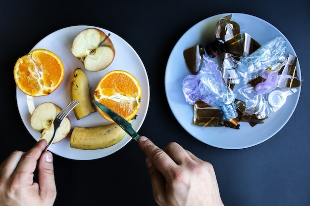 Plastic bottles and fruit on the plate. Life choice. People eating polluted food. Environmental issue. Ecological disaster. Recycling problem. Hands with knife and fork.