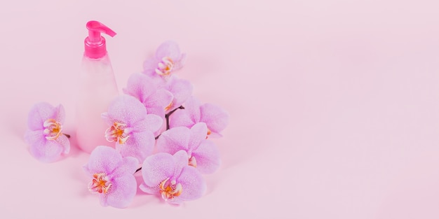 Plastic dispenser bottle with liquide cosmetic soap, intimate wash or shower gel, purple sponge and pink orchid flowers on light pink surface. Spa and women's hygiene concept