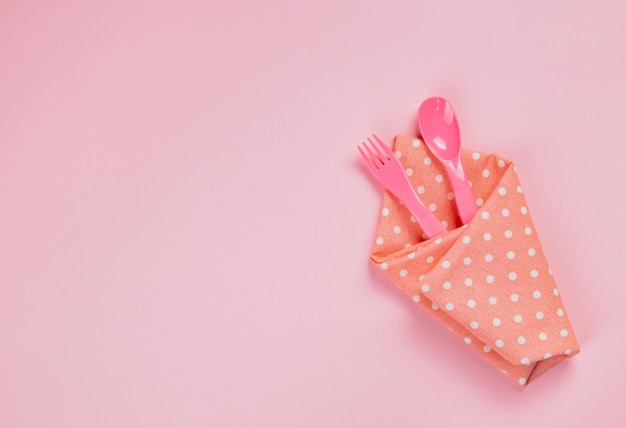 Plastic spoon, fork and pink polka dot cloth napkin on pink background