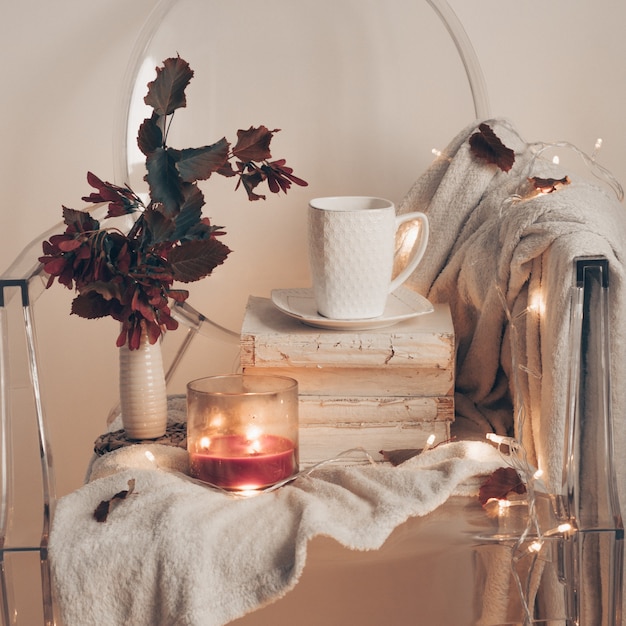 On a plastic transparent chair - Warm bedspread, a cup of tea on books and a candle with autumn leaves. Autumn winter concept.