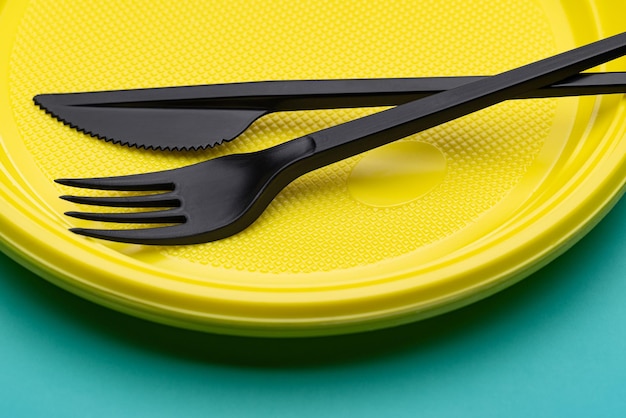 Plastic utensils close-up. Disposable yellow plate, black cutlery on a bright green background.