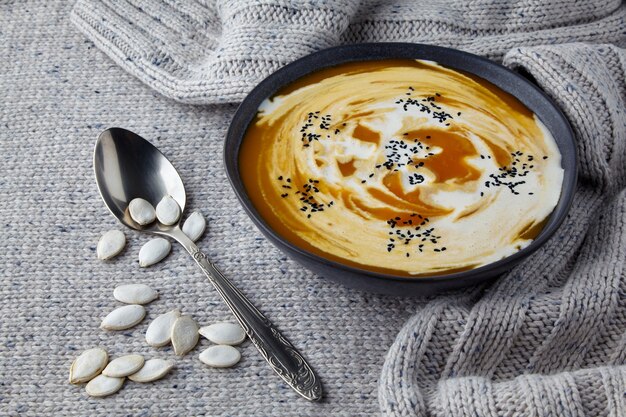 Plate of cream pumpkin soup with cream and black cumin seeds on a light knitted wool sweater. Home cooking