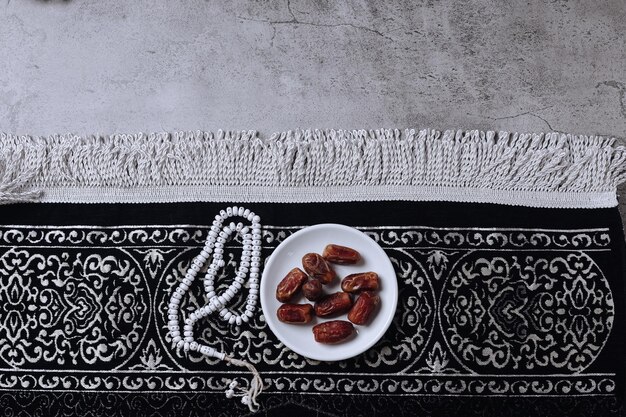 A plate of date fruits and prayer beads on prayer mat on gray background with copy space.