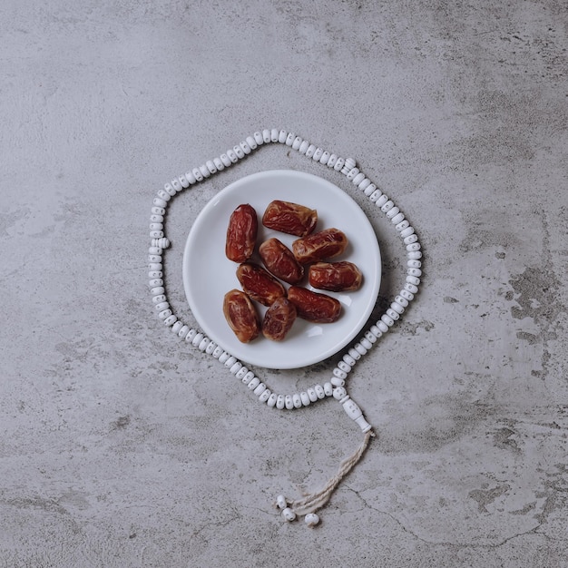 A plate of date fruits surrounded by prayer beads isolated on gray background with copy space.