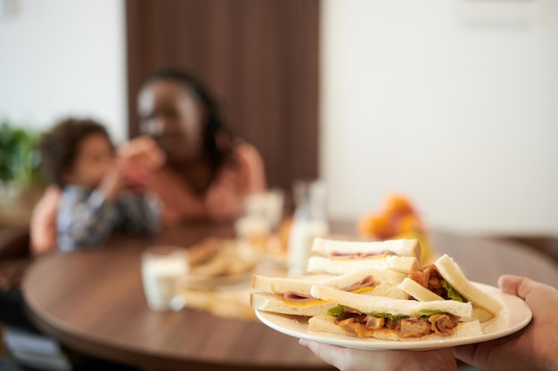 Plate of ham and chicken sandwiches in hands of woman bringing it to breakfast table