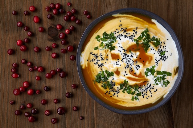 A plate of pumpkin cream soup with cream, black cumin seeds, herbs and cranberries on a wooden table. Autumn holyday