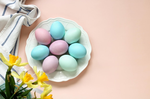 A plate with colored Easter eggs and a bouquet of daffodils on a pink background top view Flat lay