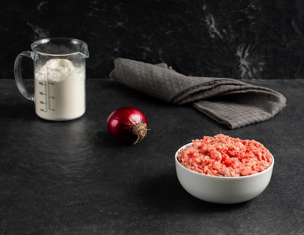 Plate with fresh ground beef red onion measuring cup with flour on a grey black background space for textxA