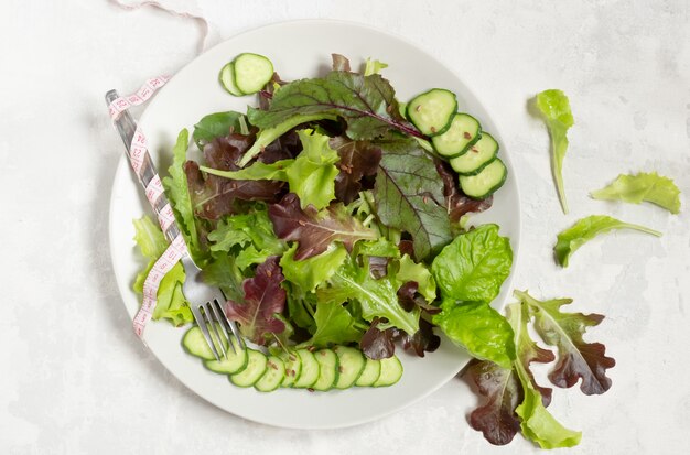 A plate with green salad leaves, sesame seeds and cucumber slices, a tape-measure wrapped around a fork on the left