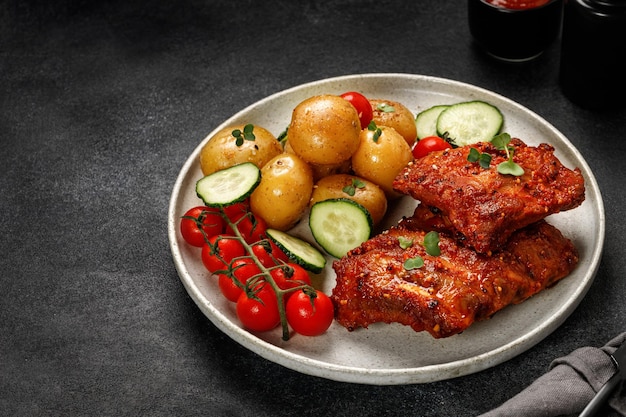 Plate with Marinated pork bbq ribs young potatoes tomatoes on black dark background Copy space