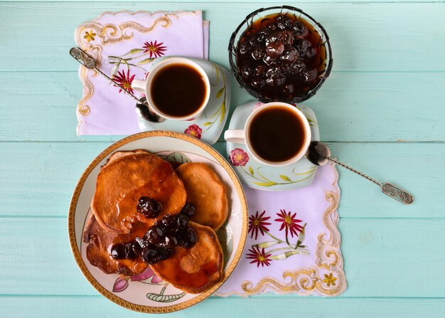 Plate with pancakes cherry jam and two cups of coffee on a wooden blue table View from above