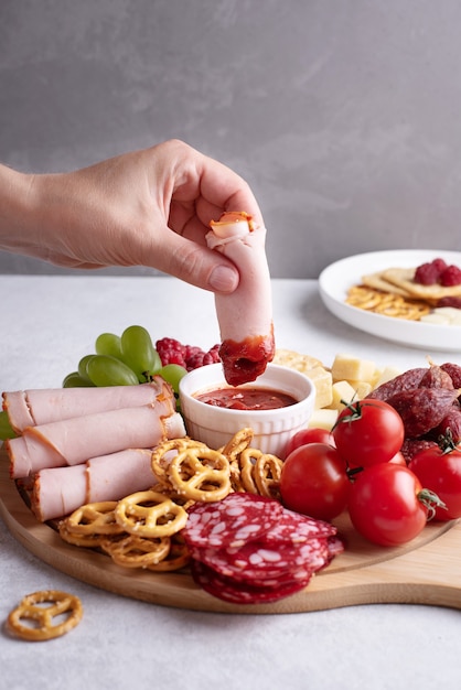 Plates with appetizer, female hand dipping a slice of ham in sauce on round charcuterie board with sausage, cheese, crackers and fruit, close-up.