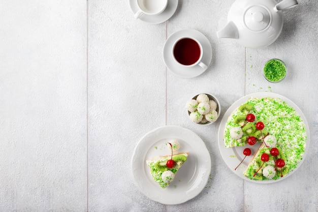 Plates with delicious cake raffaello with green coconut flakes and cup of tea on white wooden table