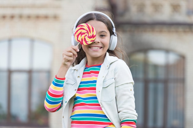 Playful beauty childhood happiness kid in digital earphones small girl listen music with lollipop back to school small girl eating sweet candy happy schoolgirl in stylish hipster outfit